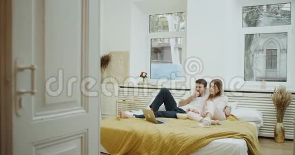 年轻有魅力的夫妇在床上吃早餐他们穿着睡衣抱着对方看着什么视频的预览图