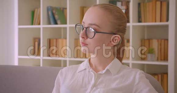年轻漂亮的白种人金发女学生戴着眼镜看着坐在沙发上的摄像机的特写照片视频的预览图