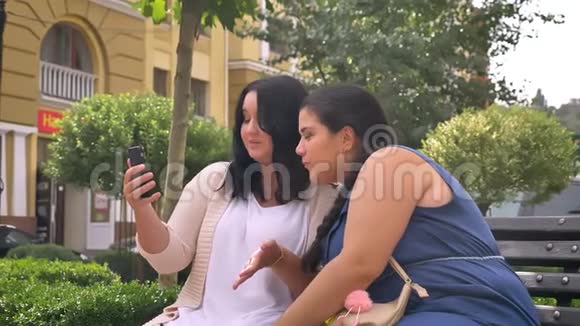 两个漂亮的白种人女孩在户外长凳上聊天聊天拿着智能手机视频的预览图