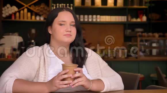 浓密可爱的白种人女性在镜头里瞥了一眼在咖啡馆里微笑着拿着纸杯视频的预览图