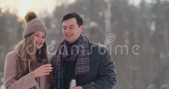 穿着外套和围巾的漂亮时髦的夫妇在冬天的森林里喝热水瓶里的茶视频的预览图