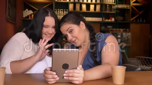 两个超重的白种人年轻女孩在一个平板电脑前挥手一边笑着坐在室内喝杯咖啡视频的预览图
