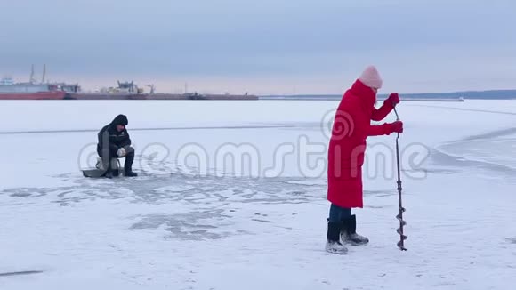 穿着红色保暖夹克和毛毡靴的漂亮渔夫们在冰冻的河流中用冰钻为冰上捕鱼打了个洞视频的预览图
