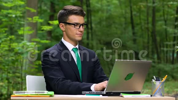 年轻微笑的上班族坐在绿色公园里的笔记本电脑上打字慢悠悠视频的预览图