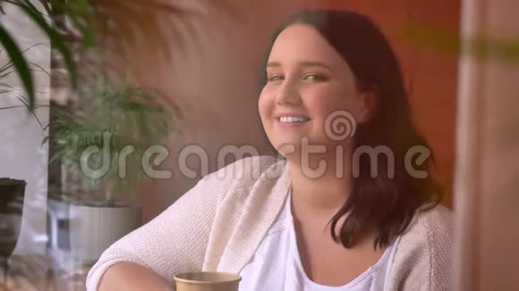 窗户附近的咖啡馆里一个大可爱的白种人女孩拿着纸杯在镜头中大笑的肖像视频的预览图
