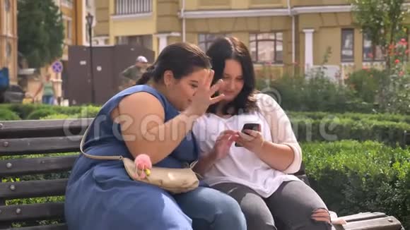 两个肥胖的白种人女孩看着智能手机在户外坐在长凳上打字视频的预览图