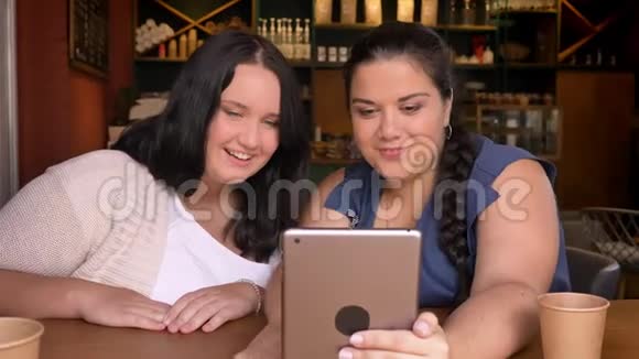 两个可爱的白种人女朋友看着同样的计划坐在咖啡馆里聊天视频的预览图