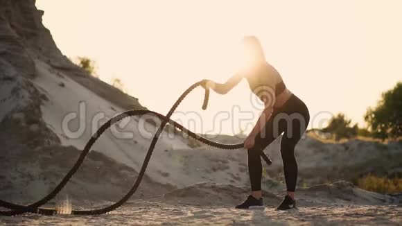 女孩运动员在日落时用绳索在海滩上训练对抗一座沙山尘土飞扬视频的预览图