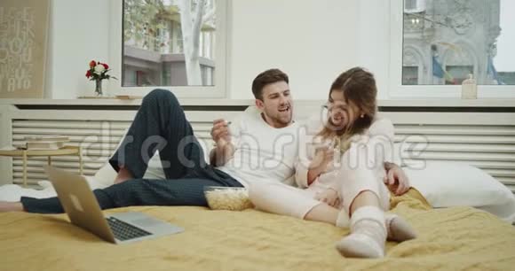 美丽的年轻女士和她的伴侣在床上穿着睡衣吃爆米花和看电影视频的预览图
