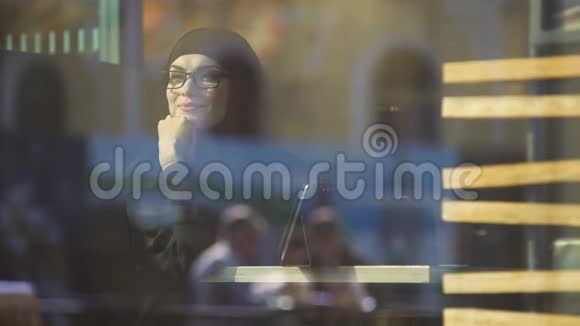 坐在靠近窗户的咖啡馆里的快乐和平的穆斯林女士看着外面自由视频的预览图