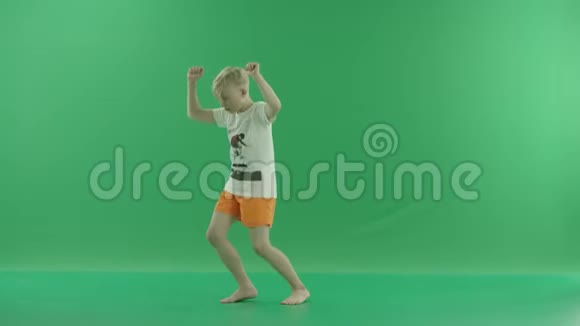 一个穿休闲服装的金发小男孩正在听音乐和跳舞他侧着身子站在绿色屏幕上视频的预览图
