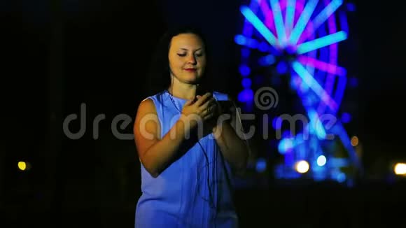一位戴着耳机的码头上的年轻女子在一个发光的摩天轮的背景下听音乐和舞蹈视频的预览图