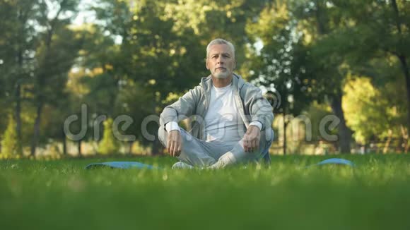 健康的老人在公园冥想坐在瑜伽垫上的莲花姿势禅宗视频的预览图