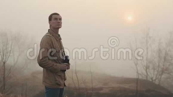 一个身穿卡其色外套的年轻红发男子手持望远镜看日出和日落四周都是雾视频的预览图