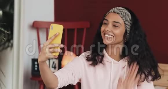 快乐的非裔美国女孩用黄色智能手机打视频电话打电话给坐在床上说话的朋友视频的预览图