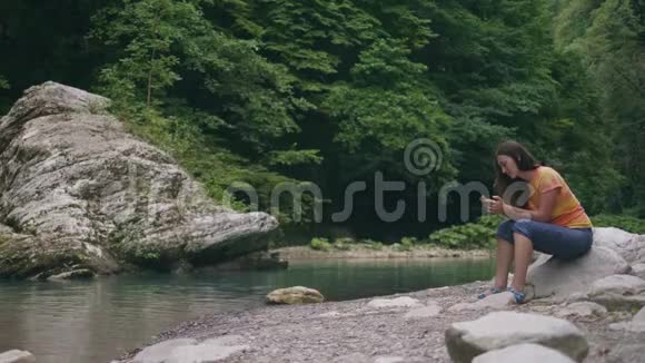江岸边休息时带手机的年轻女子野生自然景观视频的预览图
