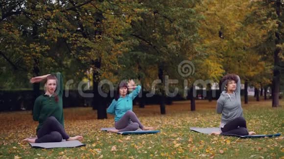 放松的女性坐在瑜伽垫上摆出牛脸姿势和伸展手臂而专业教练则在说话视频的预览图