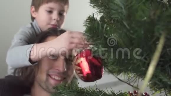 小男孩把圣诞玩具挂在树上坐在父亲的脖子上父子关系视频的预览图