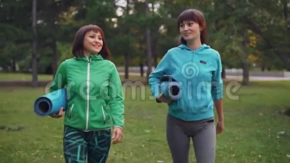 微笑的年轻女性在公园里散步他们拿着瑜伽垫聊天分享新闻一起讨论练习青年视频的预览图