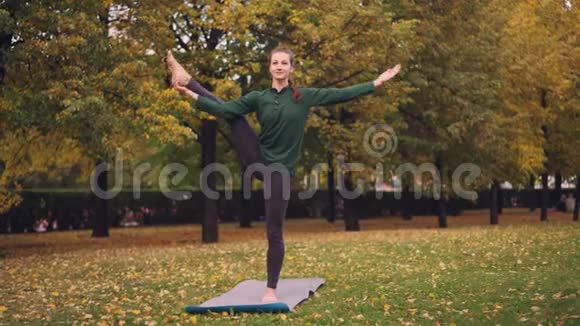 微笑女孩职业瑜伽教练正在做平衡练习站在草地上的一条腿上视频的预览图