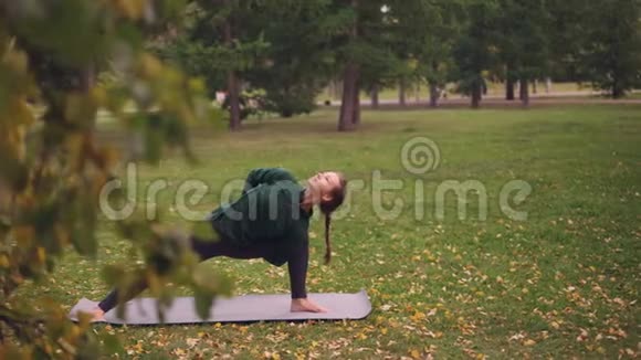 瑜伽学生正在公园练习瑜伽垫上的侧角位置Parsvakonasana的变体绿和黄树视频的预览图
