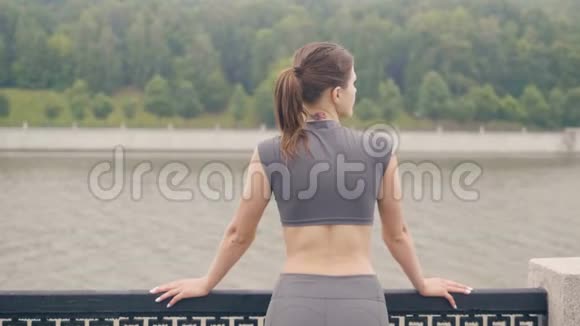 穿着运动服的年轻女子站在夏季公园的河堤上视频的预览图