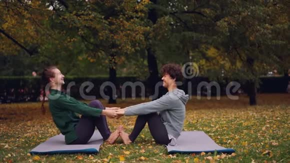 快乐的年轻女性正在做一对瑜伽玩得开心坐在公园草地上的垫子上笑美丽的秋天视频的预览图
