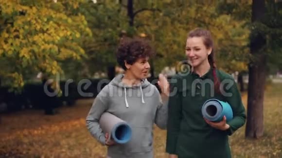 美丽的女孩正在公园里散步瑜伽教练拿着垫子说话手势和笑声师生视频的预览图