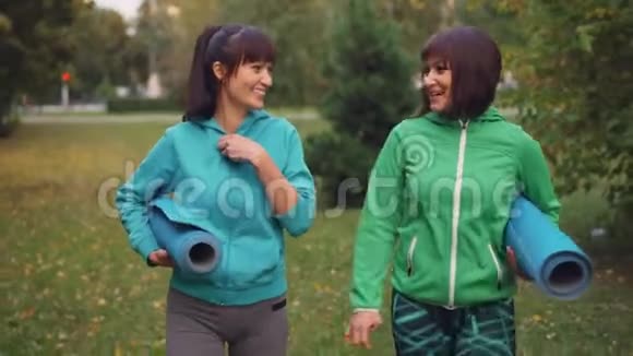 多莉拍摄的漂亮女运动员交谈微笑和拿着瑜伽垫在公园散步在秋天的一天城市视频的预览图