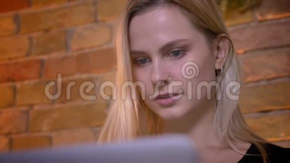 年轻漂亮金发女千禧一代在笔记本电脑上打字坐在室内沙发上的特写照片视频的预览图