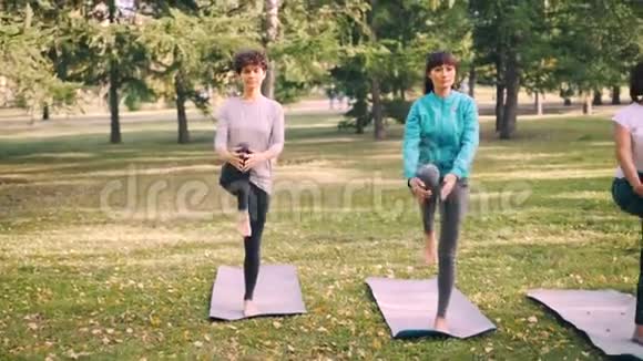 女瑜伽学生在公园的垫子上站在一条腿上做平衡运动女孩在训练中接受指导视频的预览图