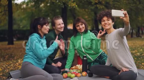 有魅力的年轻女士瑜伽迷在秋天在公园野餐时使用智能手机自拍女孩们摆姿势视频的预览图