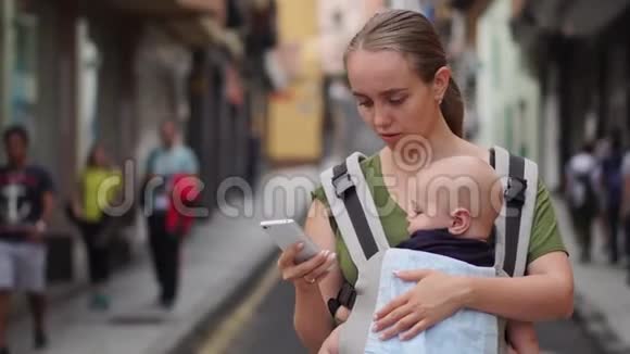 妈妈站在街上带着一个穿着吊带的孩子看着手机屏幕视频的预览图