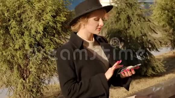 漂亮的金发女孩戴着黑色外套帽子在阳光明媚的日子里坐在植物旁边的现代背包里拿出手机视频的预览图