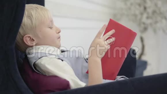 一个严肃的金发男孩坐在椅子上读一本厚厚的书视频的预览图