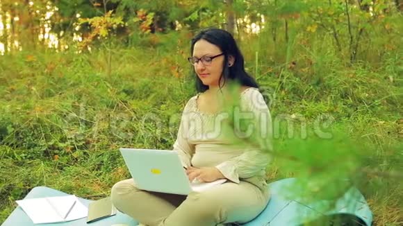 一个带眼镜的黑发女人戴着眼镜在林中空地上用笔记本电脑工作视频的预览图