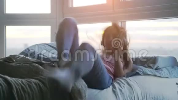 在日落时分戴着耳机的年轻漂亮快乐的女人躺在靠窗的床上戴着镜片闪光效果和视频的预览图