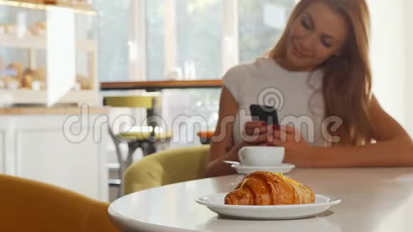 有选择地关注美味的羊角面包在咖啡店使用智能手机的女人视频的预览图