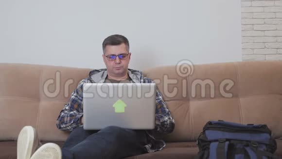 一位坐在沙发上的中年男子使用笔记本电脑旁边是一个大型旅游背包视频的预览图