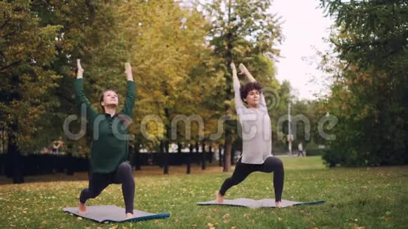 微笑的女孩瑜伽老师和学生在户外的草地上锻炼在绿色的垫子上练习瑜伽视频的预览图