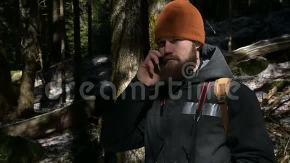 一个戴着帽子的长胡子的人在针叶林里用手机说话细胞通讯的概念视频的预览图