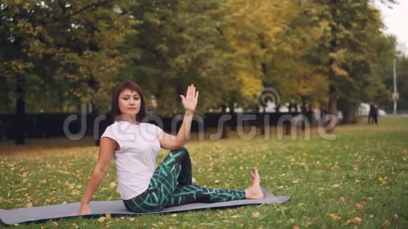 穿着运动服的漂亮女孩正在公园里锻炼身体在温暖的秋天练习坐在瑜伽垫上的弯曲姿势视频的预览图