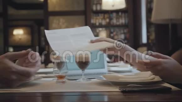 无法辨认的男人和女人坐在咖啡馆的桌子旁工作笔记本电脑文件和带咖啡的眼镜都在上面视频的预览图