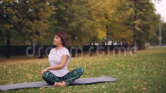 柔柔女瑜伽尼正在练习简单的扭转姿势然后坐在公园草地上的瑜伽垫上以莲花的姿势休息视频的预览图