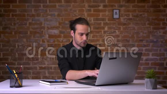英俊的白种人坐在笔记本电脑前在靠近砖墙的办公室里打字视频的预览图