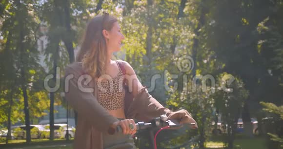 在公园里年轻漂亮的白种人女性站着踢滑板车看着摄像机视频的预览图
