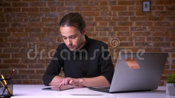 可爱的白种人一边坐在他的工作地点一边在后面的砖墙上滚动着他的平板电脑视频的预览图