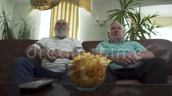 两个成熟的高级男人坐在棕色的皮革沙发上看电视朋友同时向前倾等待视频的预览图