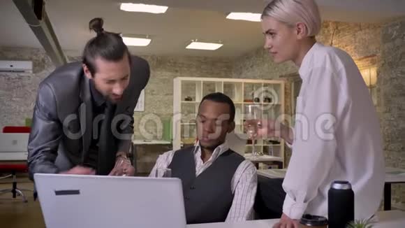 三个不同种族的工人讲故事坐在办公室的笔记本电脑旁喝香槟交流放松视频的预览图