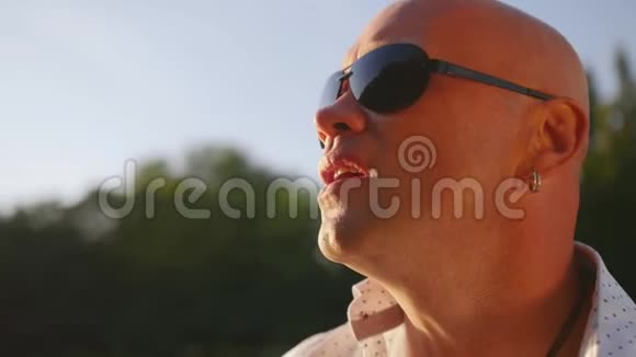 一个戴太阳镜的秃头成年男子在公园户外唱歌的肖像近距离射击视频的预览图